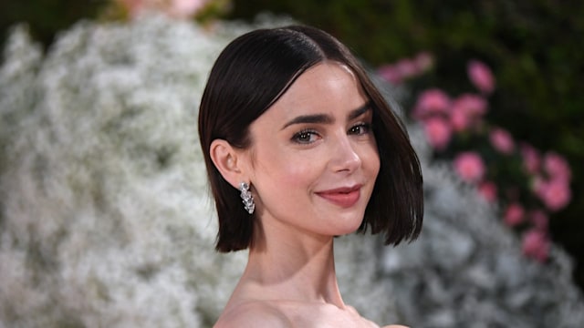 The image shows actress Lily Collins with a sleek, shoulder-length bob hairstyle, parted in the middle. She is wearing diamond drop earrings and smiling softly. Her makeup is natural with a hint of blush and light pink lipstick. The background features soft, blurred greenery and pink flowers, enhancing her elegant appearance.