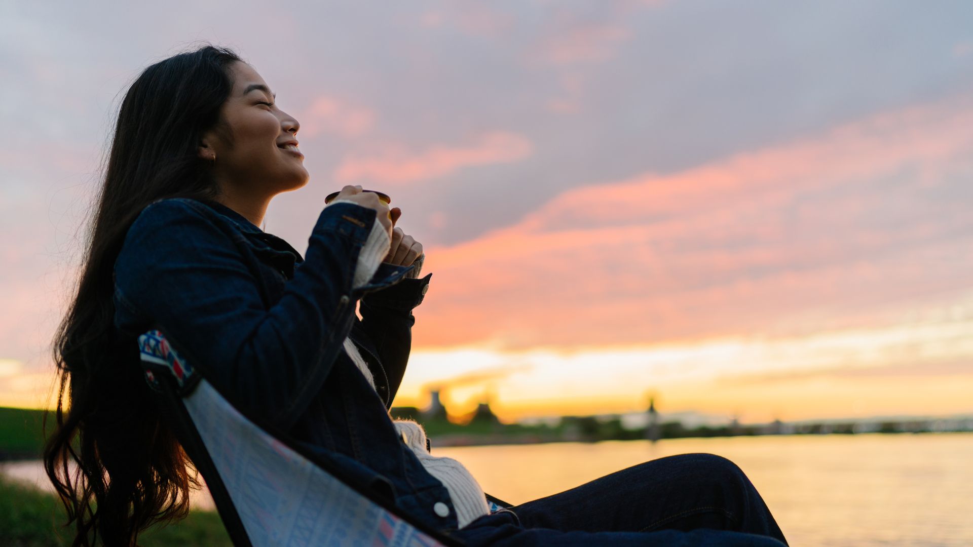 A young woman is enjoying sitting near a lake and drinking a hot drink in nature during sunset.