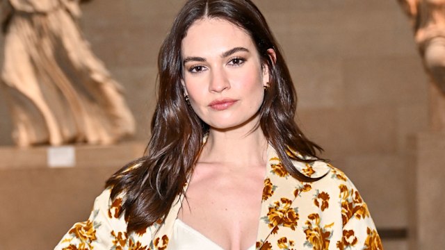 The image shows Lily James wearing a chic matching set featuring a beige fabric adorned with bold golden-yellow floral patterns. Her outfit includes a crop top and a skirt with a blazer. She stands confidently with minimal makeup, sleek loose waves, and subtle accessories, exuding elegance. The background features classical statues.