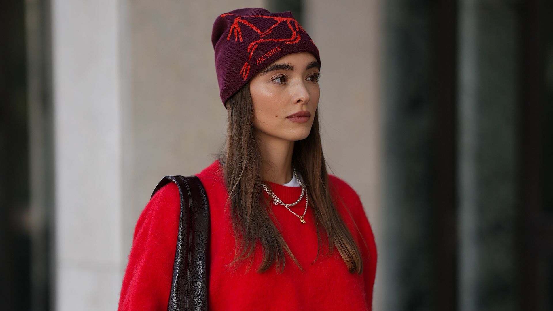 Evi Wave is seen wearing a bright red knit sweater with long sleeves from American Vintage; a white long-sleeved t-shirt, worn under the sweater from Weekday; wide-leg, brown trousers with a voluminous silhouette from COS; a large bordeaux leather shoulder bag with a glossy shade from Arket; subtle silver jewellry, featuring metallic necklaces and rings from Ariane Ernst