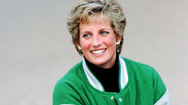 Princess Diana leans on a fence, sporting the green Eagles jacket with a big smile. With tousled hair and gold accessories, she turns casual sportswear into a fashion statement.
