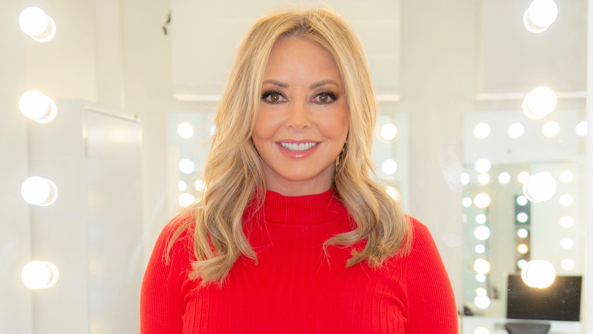 Carol Vorderman sitting on a desk in red outfit
