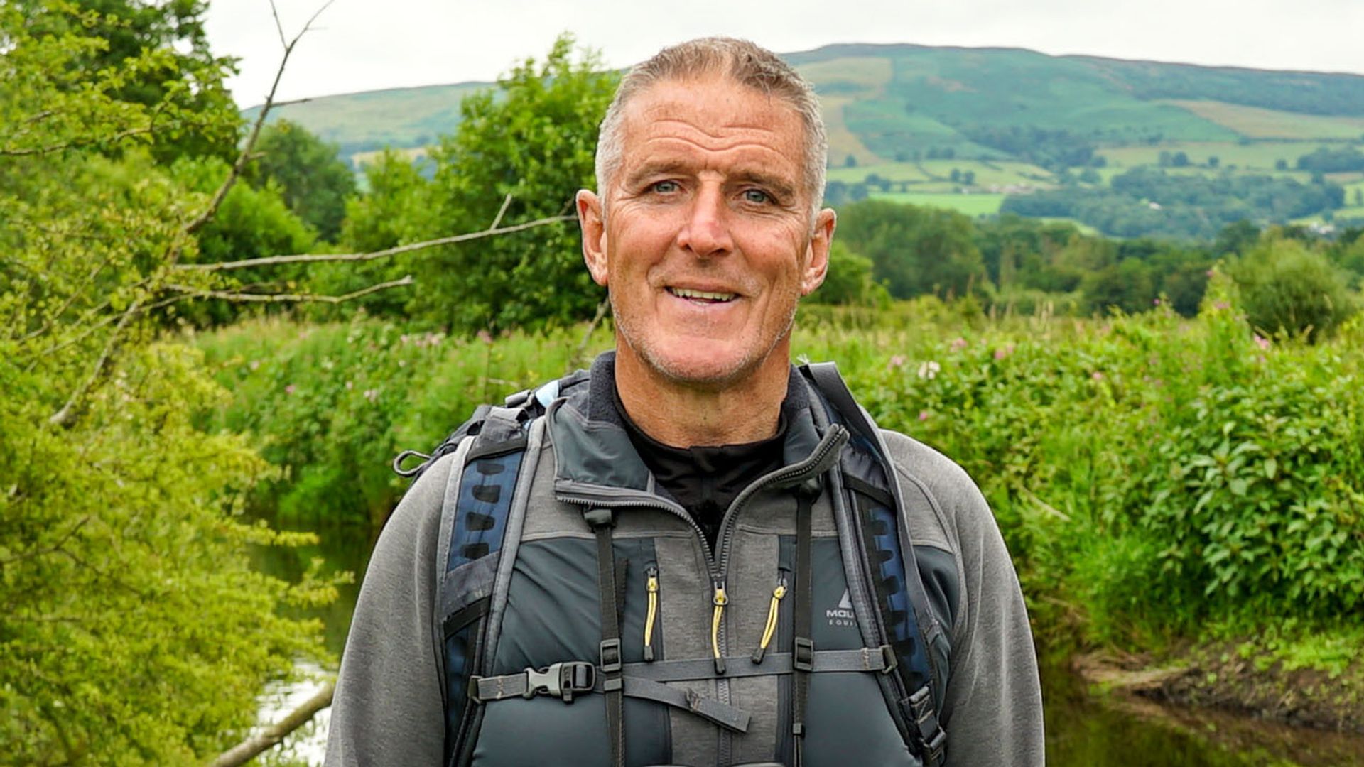 Iolo Williams in a grey wetsuit standing by a river