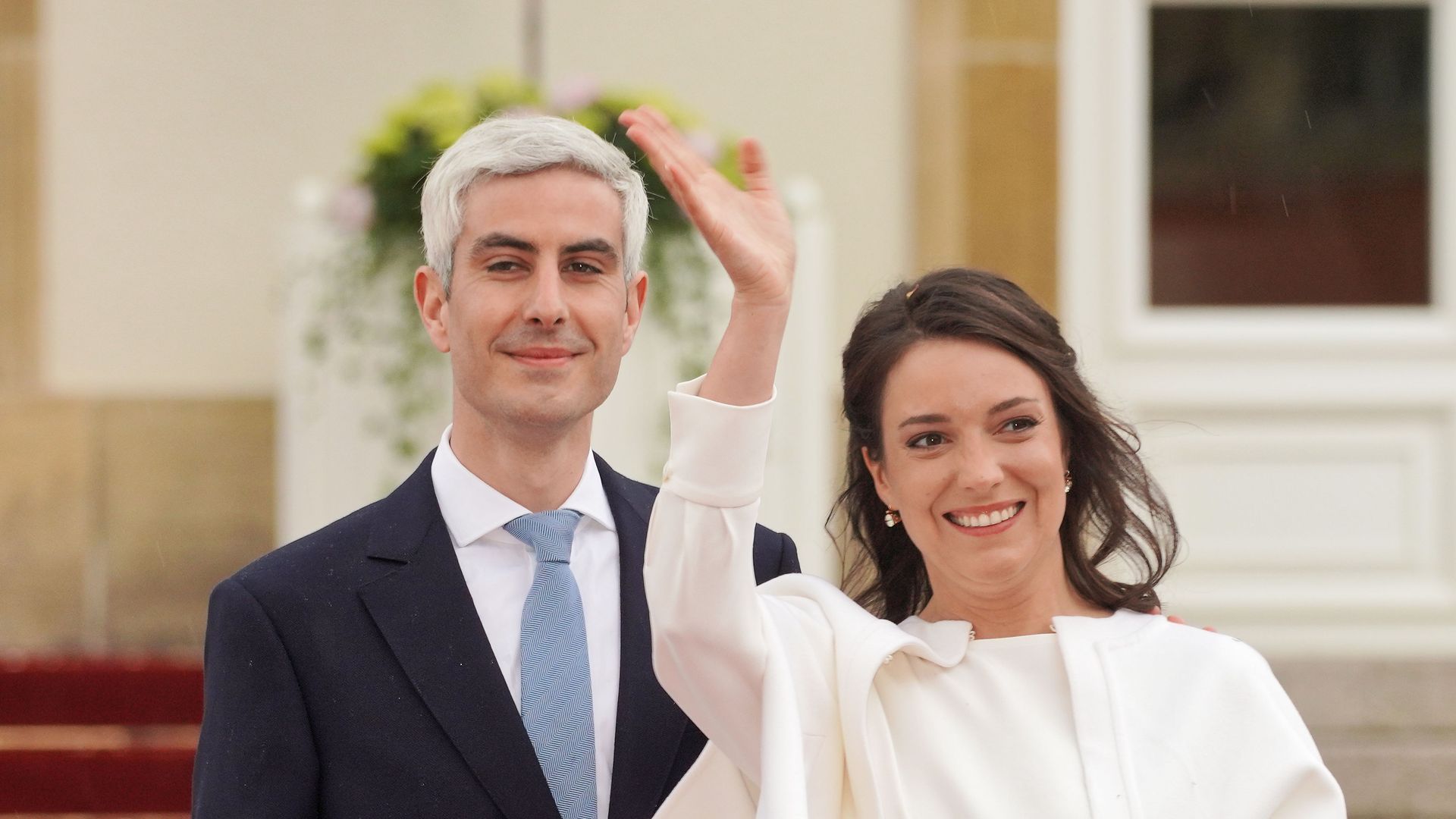 woman in white suit waving joined by man in suit 