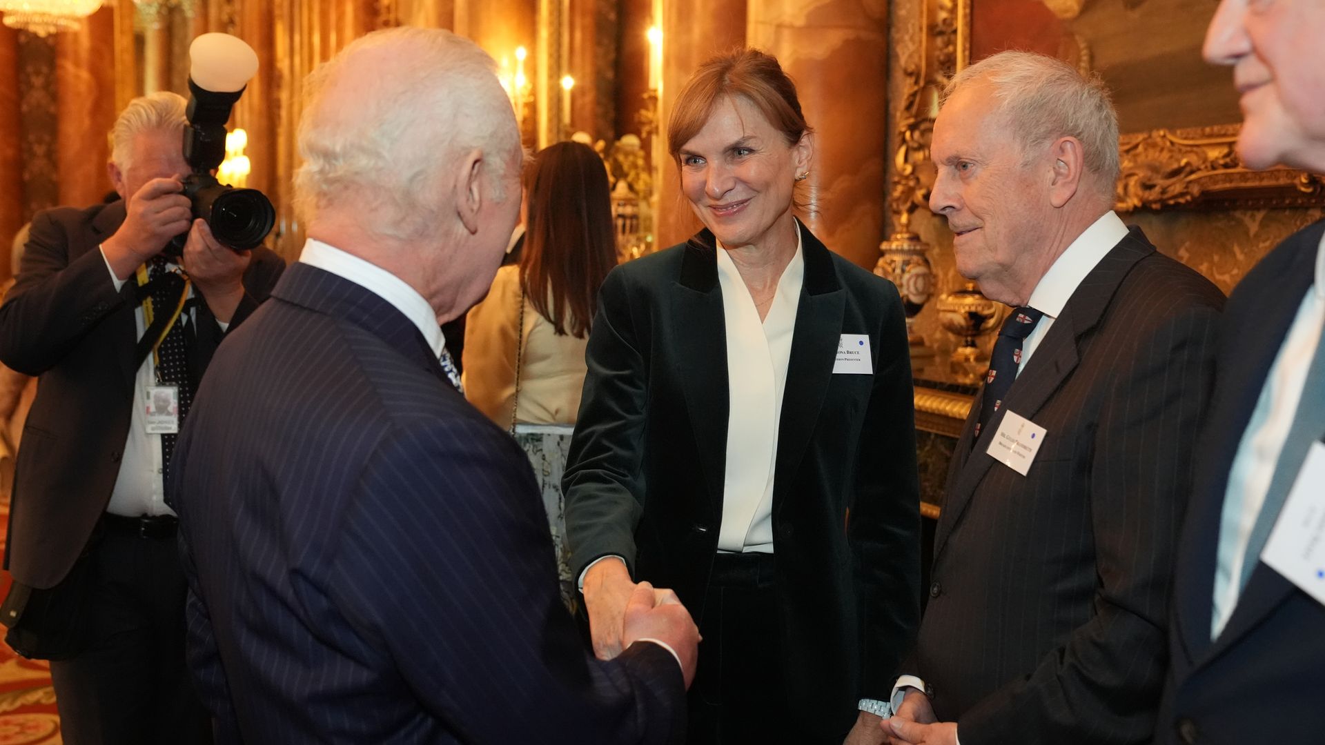 King Charles shaking hands with Fiona Bruce, standing next to them is Gyles Brandreth