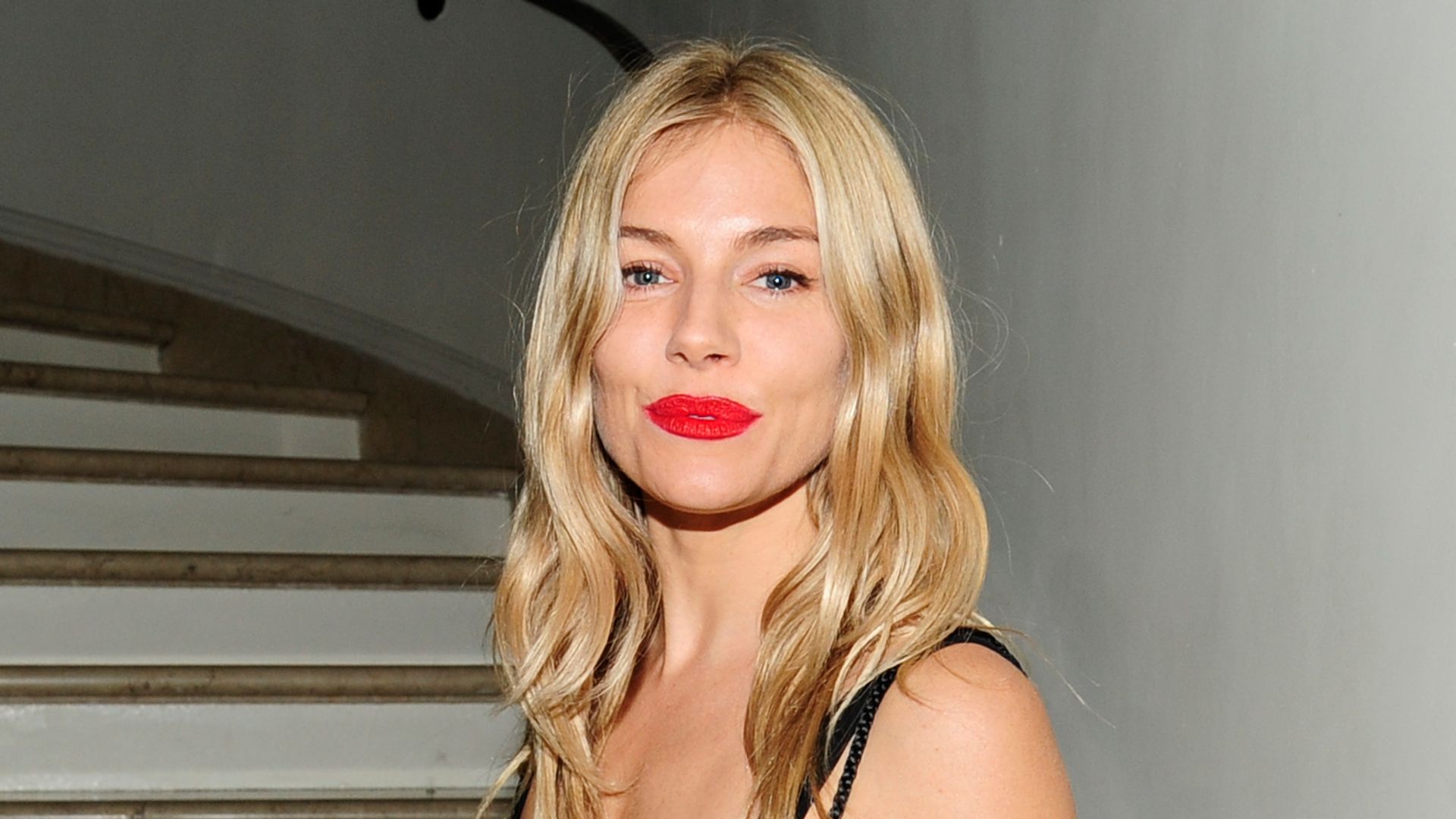 Sienna Miller smiling in a floral dress with red lipstick and flyaway hair 