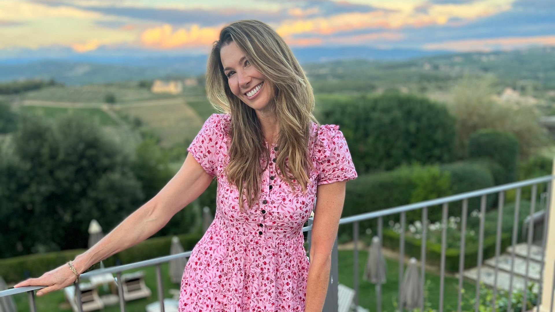Rosie Green smiling on a balcony in a pink floral dress