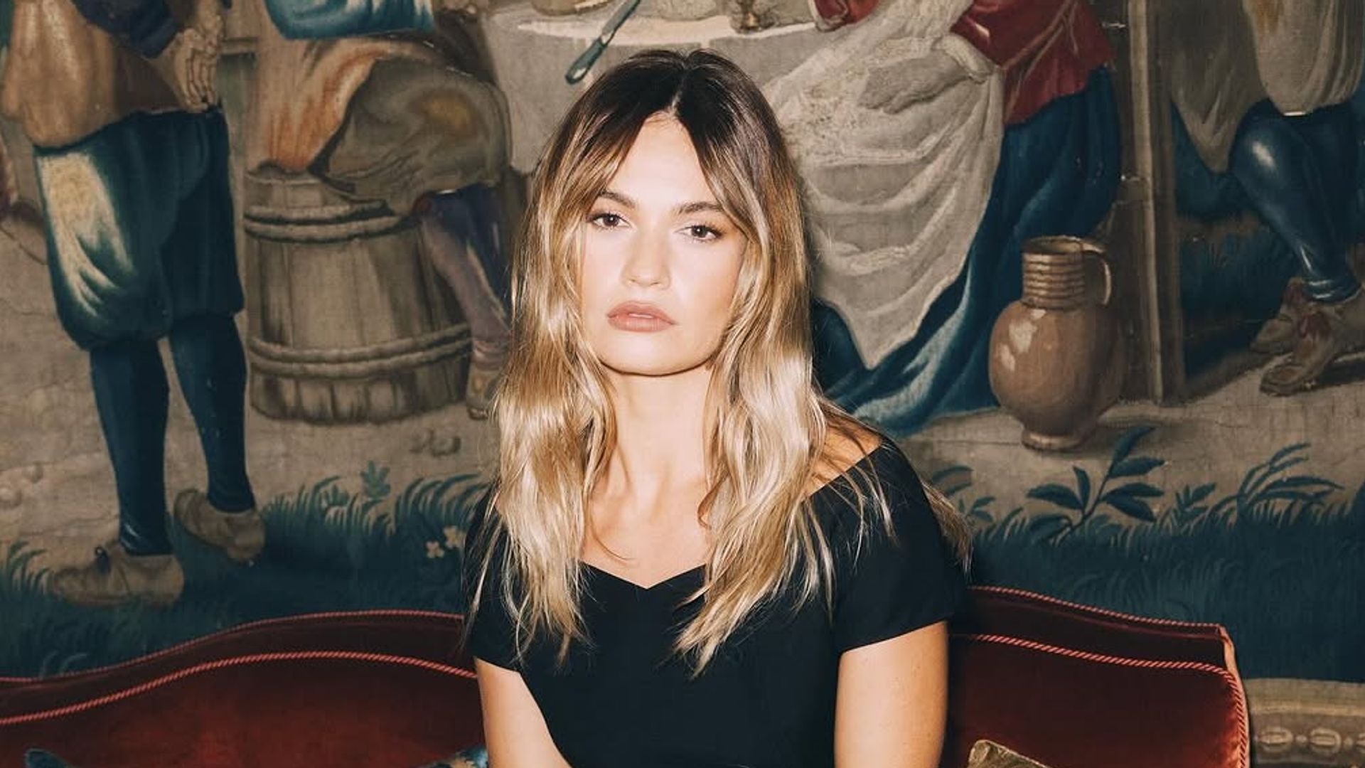 Lily James poses inside a Parisian salon wearing an off-the-shoulder black Dior look, seated on a plush red velvet sofa with a Lady Dior handbag beside her. A historical tapestry serves as an opulent, storybook-like backdrop.