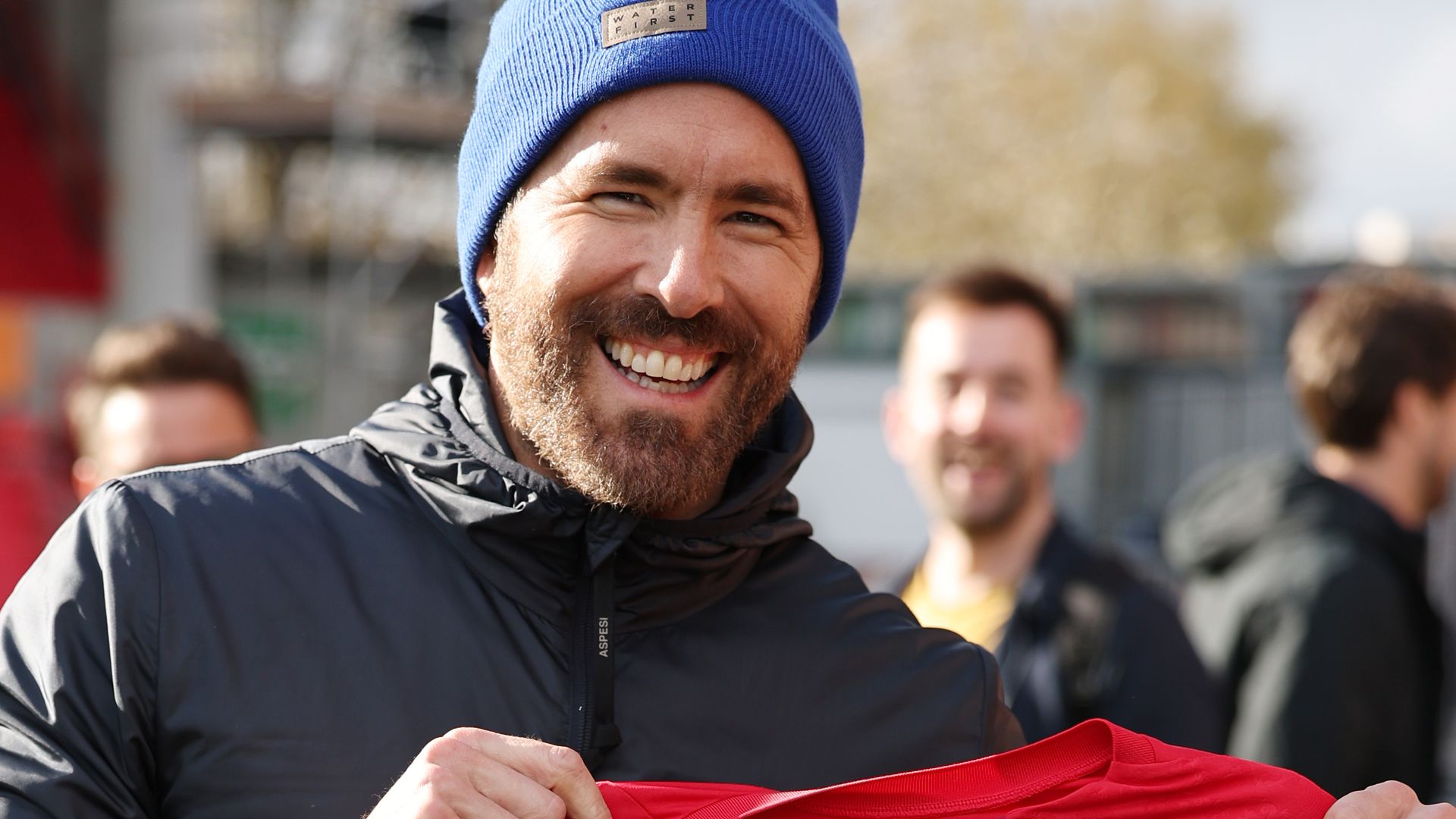 Ryan Reynolds smiling holding up a red football shirt