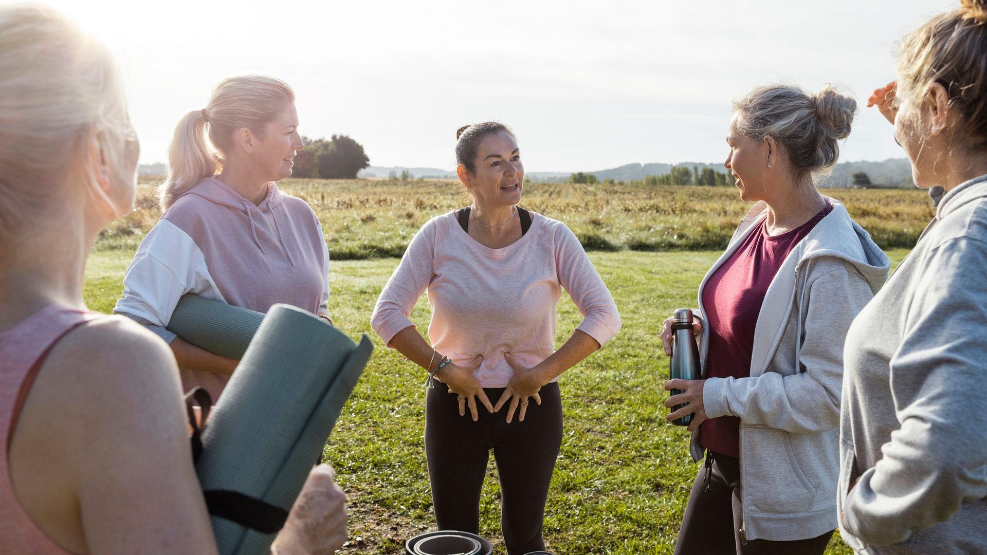 Group of mature women doing Yoga outdoors in France together while on a wellness vacation. They are all standing holding their mats ready to get started. The yoga instructor is giving advice on the exercises they will be doing.