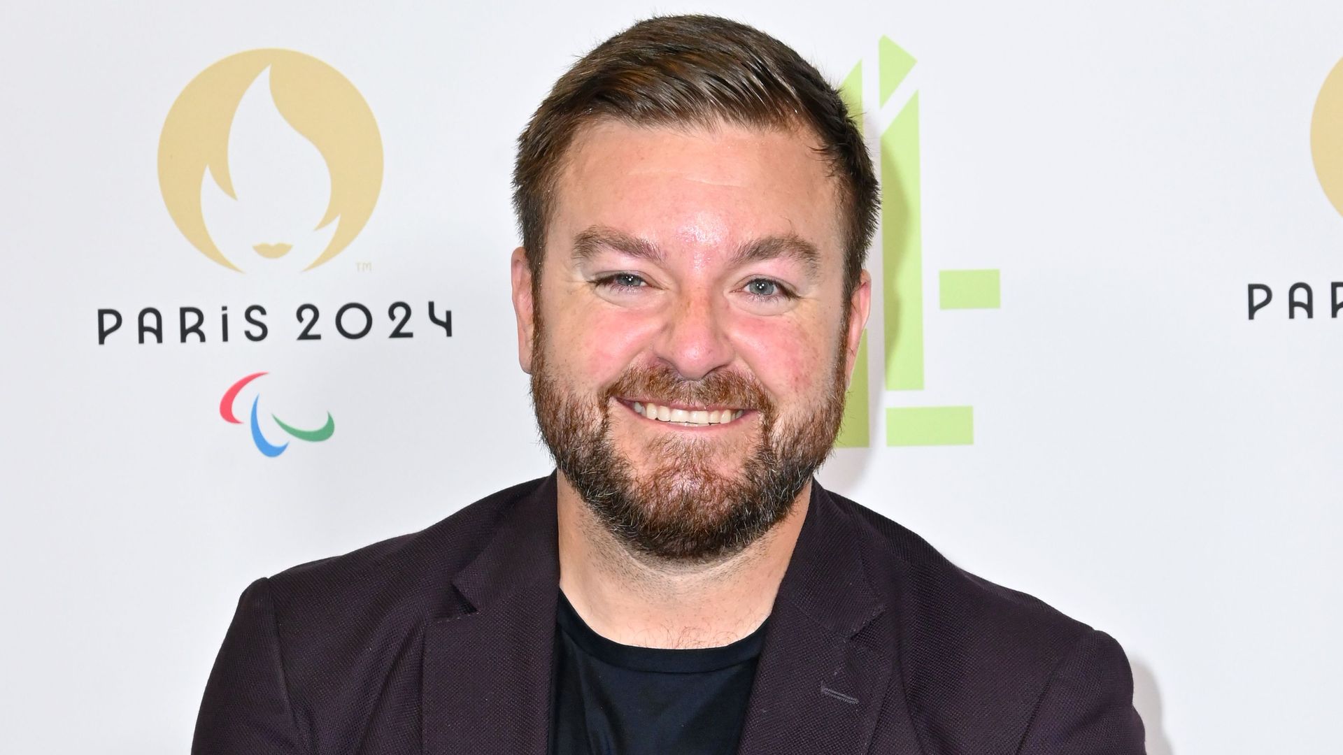 Alex Brooker standing in a dark suit in front of photographers board at Paralympic Games Paris 2024 launch