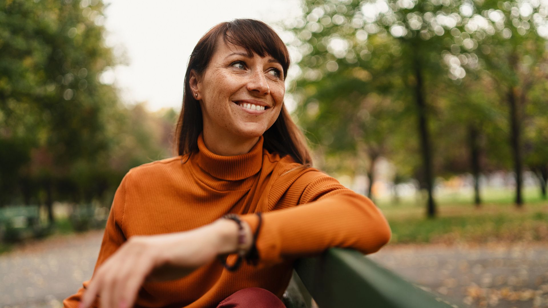 Happy female in turtleneck with freckles sitting outdoors in park.