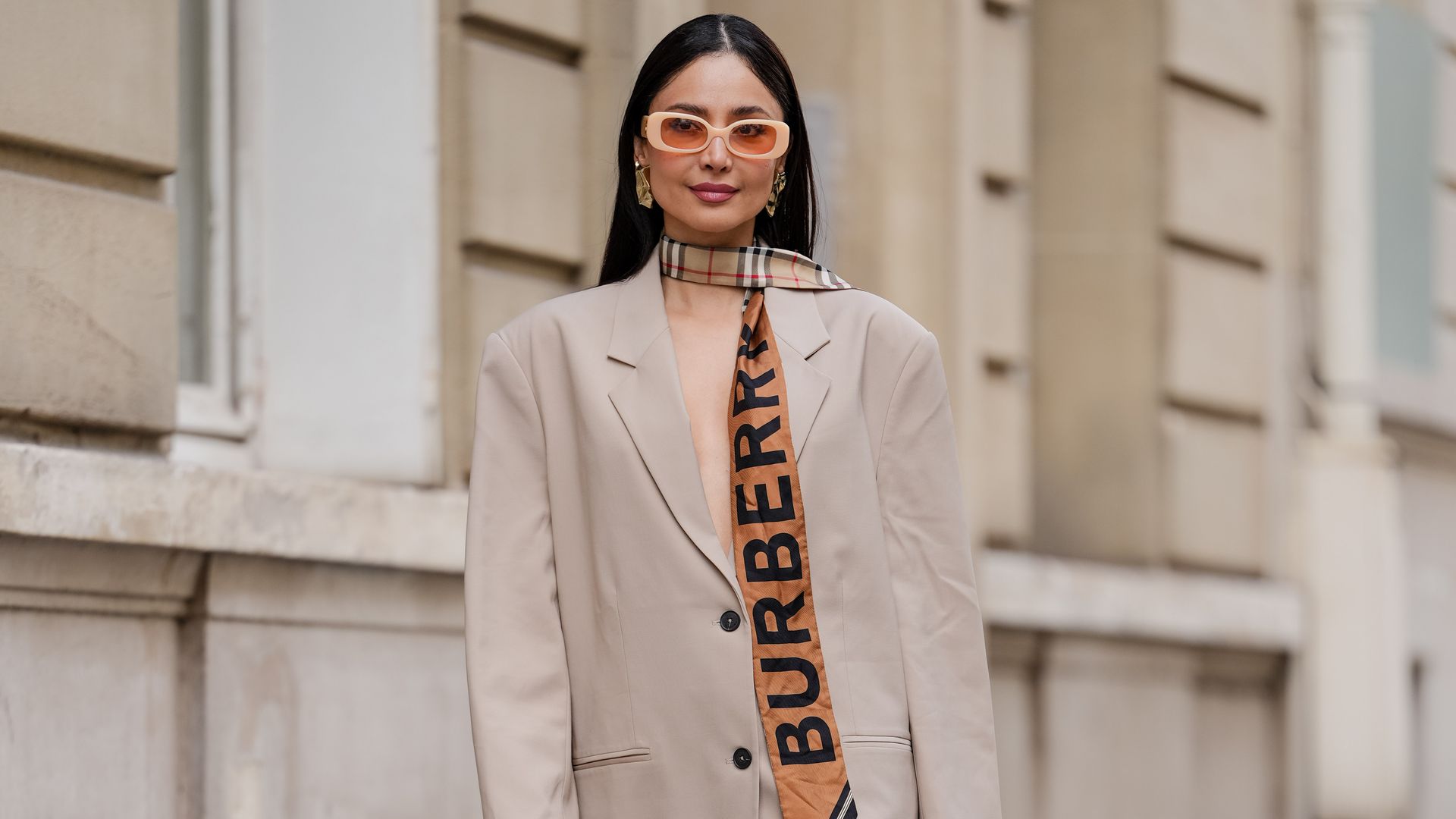 atricia Gloria Contreras wears long, straight black hair with a middle part, dark lipstick and defined brows. Oversized square sunglasses by Linda Farrow are styled with sculptural gold earrings and a beige Burberry scarf with bold logo lettering tied around the neck. 
