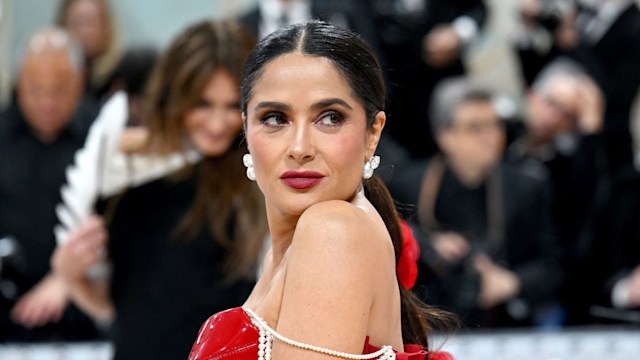 Salma Hayek poses at the Met Gala wearing a bold red latex gown with pearl shoulder draping, statement earrings, sleek hair, and bold makeup, standing out against a blurred crowd backdrop.