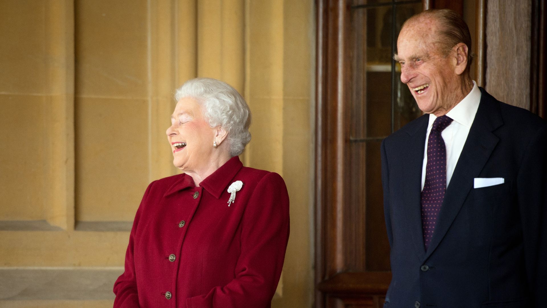 Queen Elizabeth II laughing whilst wearing a red coat and Prince Philip smiling in a blue suit