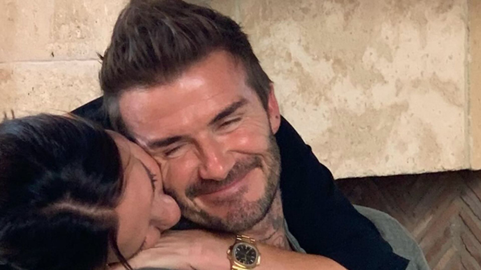 Victoria Beckham kissing the side of David Beckham's face with her arm draped over him