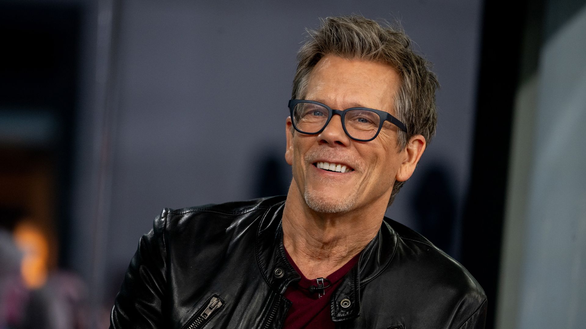 Kevin Bacon smiles wearing leather jacket and glasses