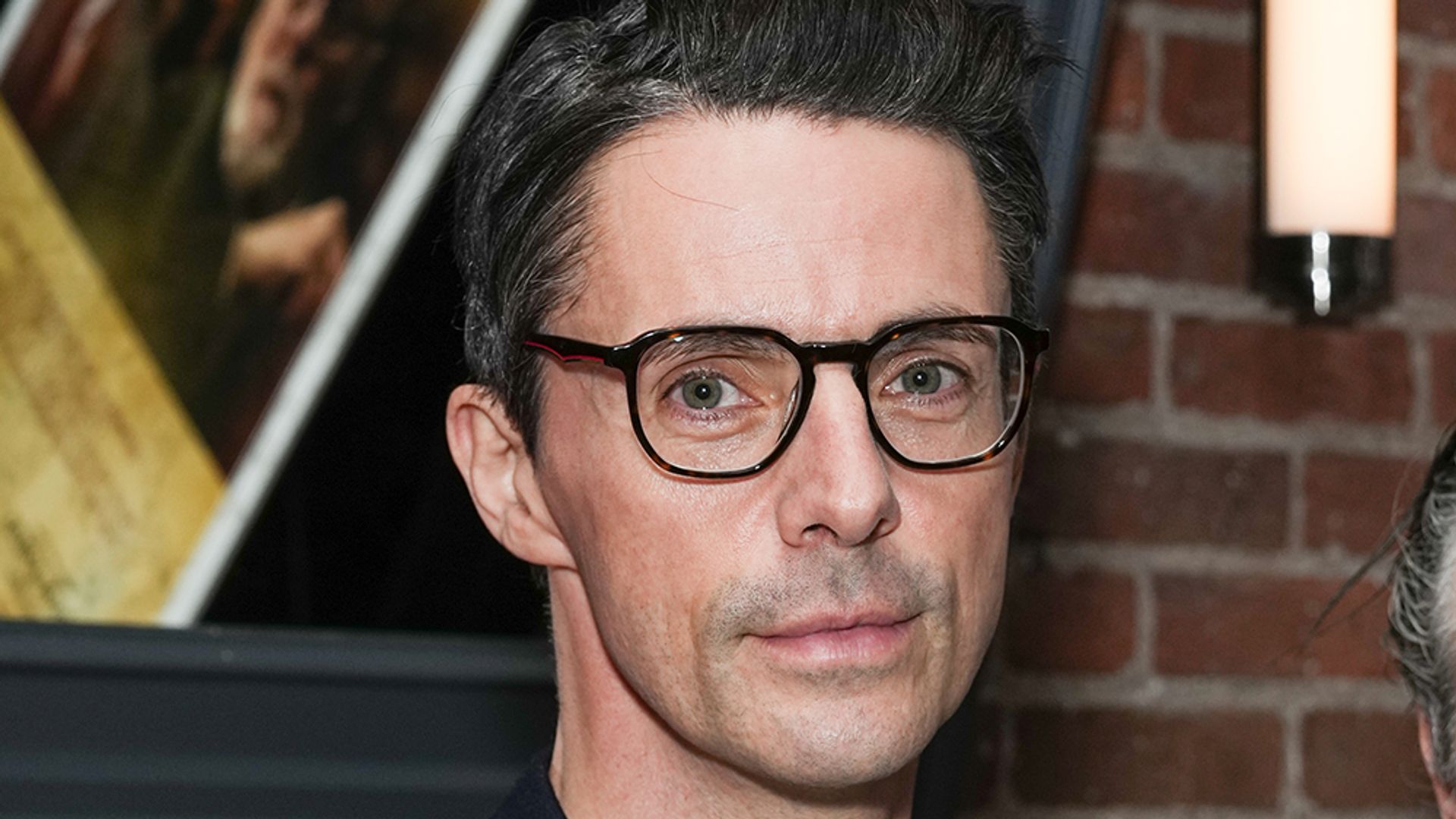 Matthew Goode in a black suit and glasses 