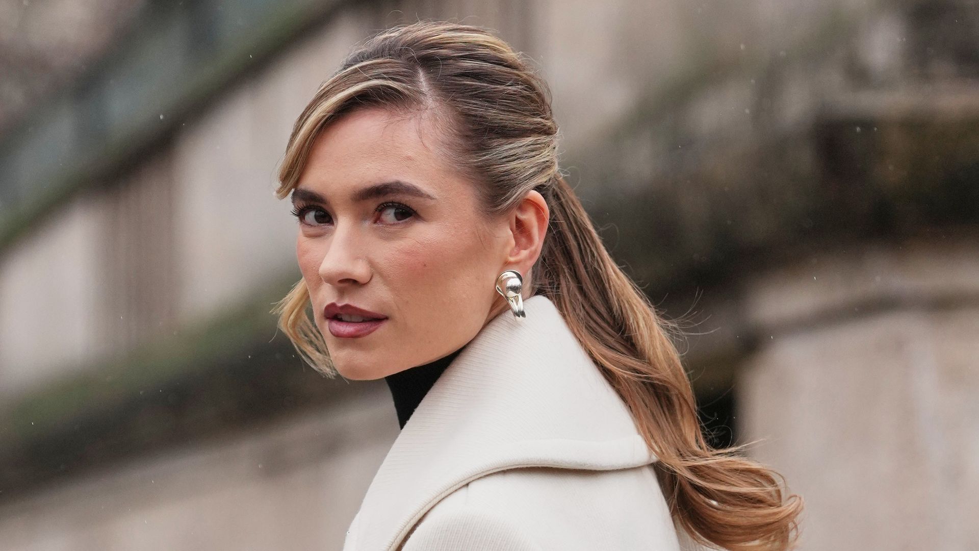Zita D'hauteville wears blonde shoulder length hair with a side part styled in loose waves ponytail, metal hoop earrings, a brown leather shoulder bag, a white wool coat with wide notch lapels and silver tone double breasted buttons and a black "PATOU" sleeve patch, a black rib knit turtleneck top, outside Patou, during Paris Fashion Week - Menswear Fall/Winter 2026-2027, on January 25, 2026 in Paris, France (Photo by Edward Berthelot/Getty Images)