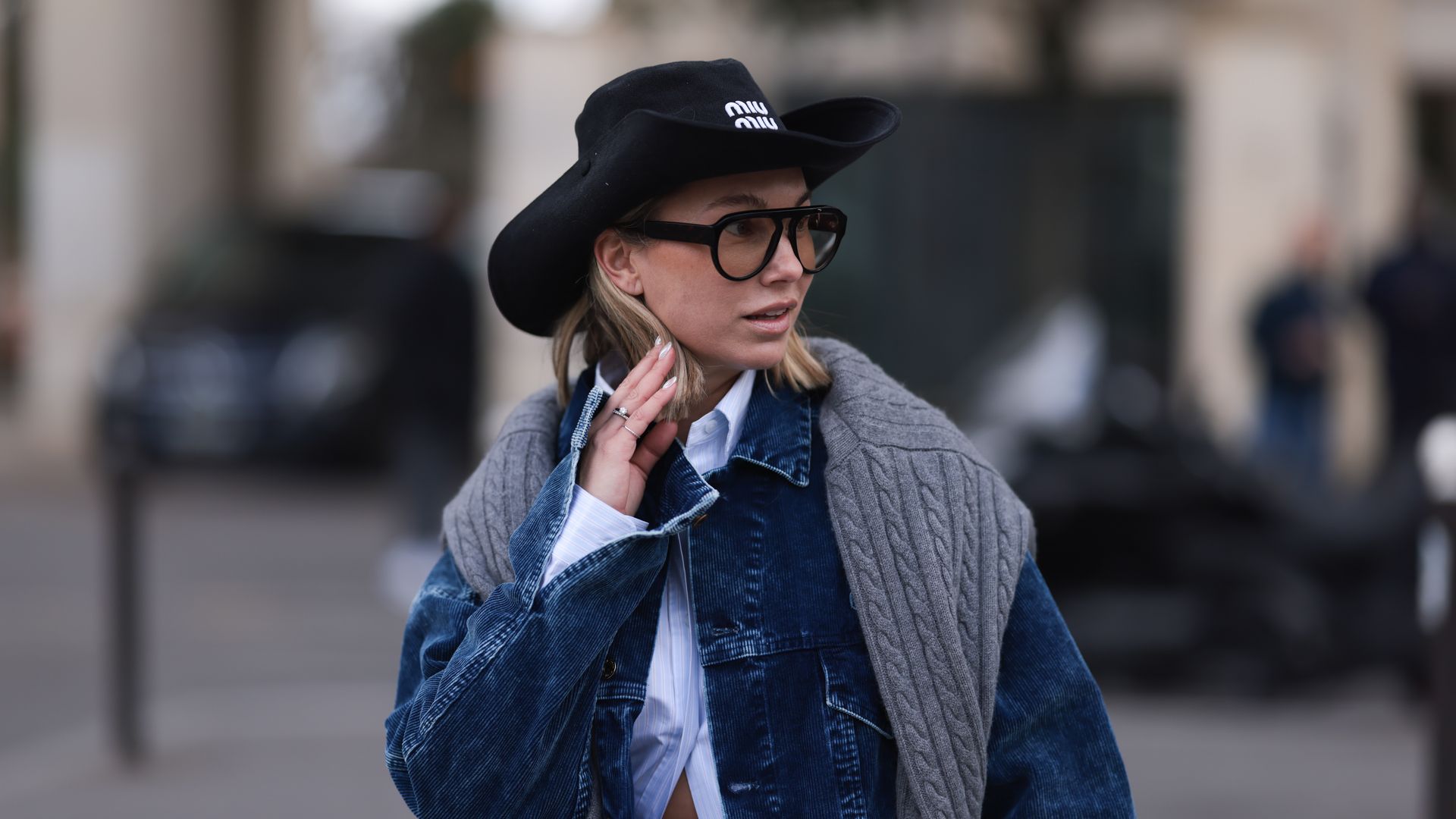 Karin Teigl seen wearing Miu Miu black denim logo cowboy hat, black aviator glasses, Miu Miu light blue striped buttoned shirt, Miu Miu grey cable wool knit sweater, Miu Miu blue corduroy jacket, before Miu Miu, during the Womenswear Fall/Winter 2024/2025 as part of Paris Fashion Week on March 05, 2024 in Paris, France.
