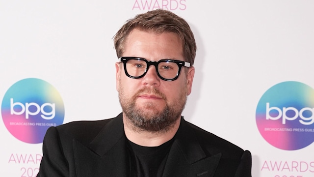 James Corden in a black suit clasping his hands together while standing in front of BBC Studios step and repeat board