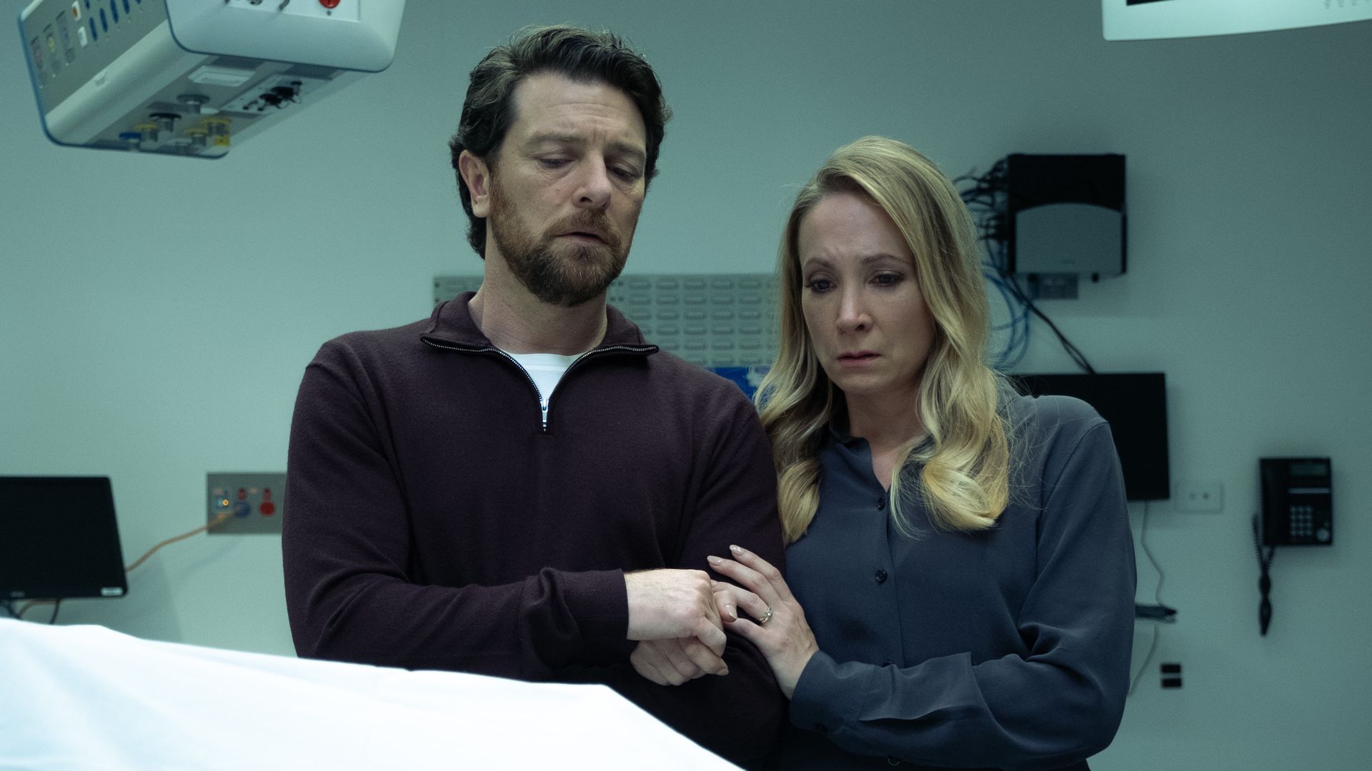 husband and wife standing over covered body in morgue