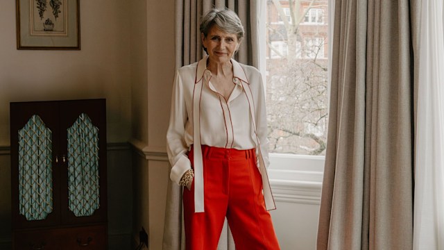 A woman standing next to a window whilst wearing red trousers and a white blouse