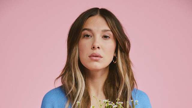 Millie Bobby Brown poses infront of a pink backdrop holding flowers