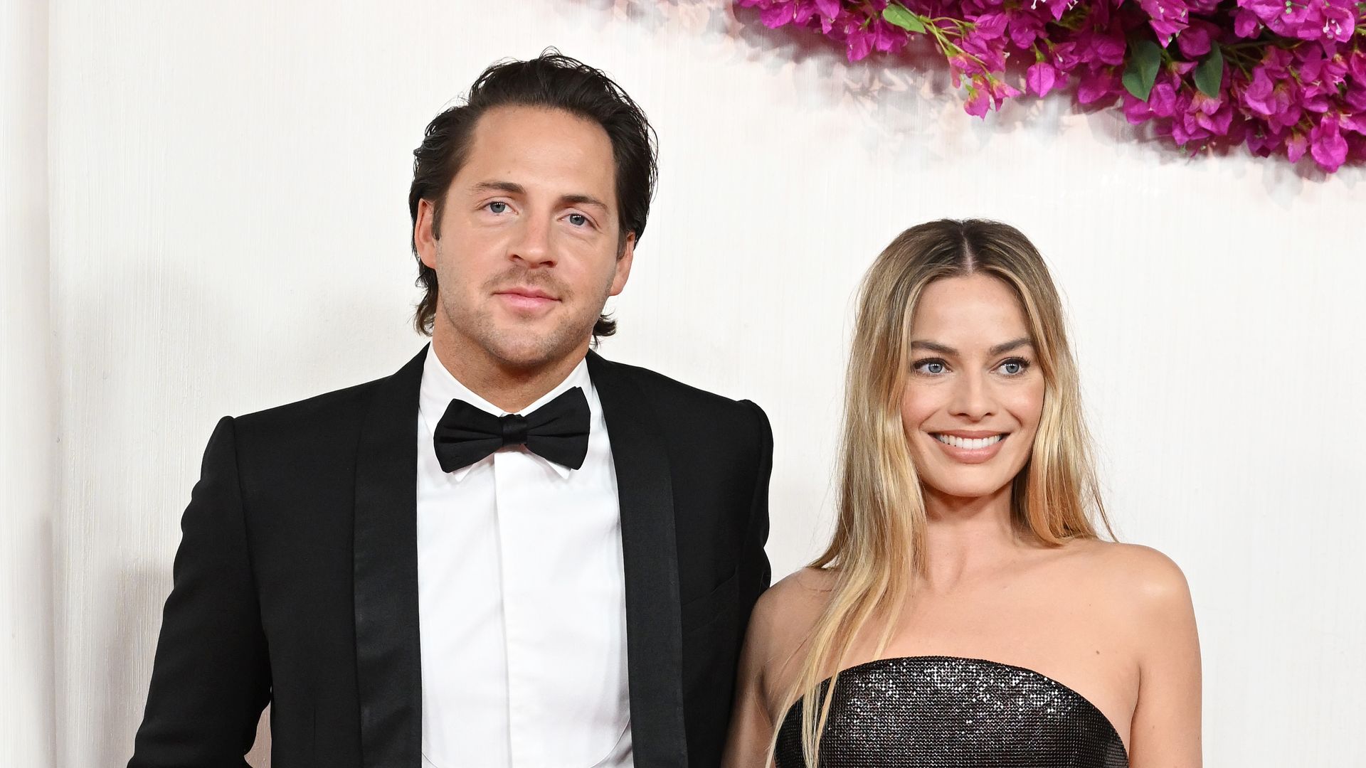 Tom Ackerley and Margot Robbie smile for photographers in front of white screen while attending Oscars