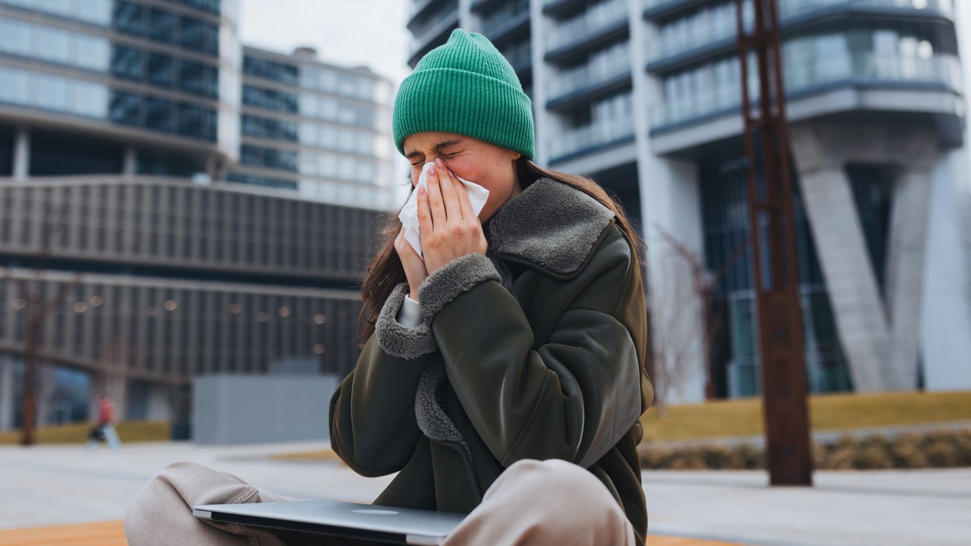 Sick university student on bench in front of university building blowing nose