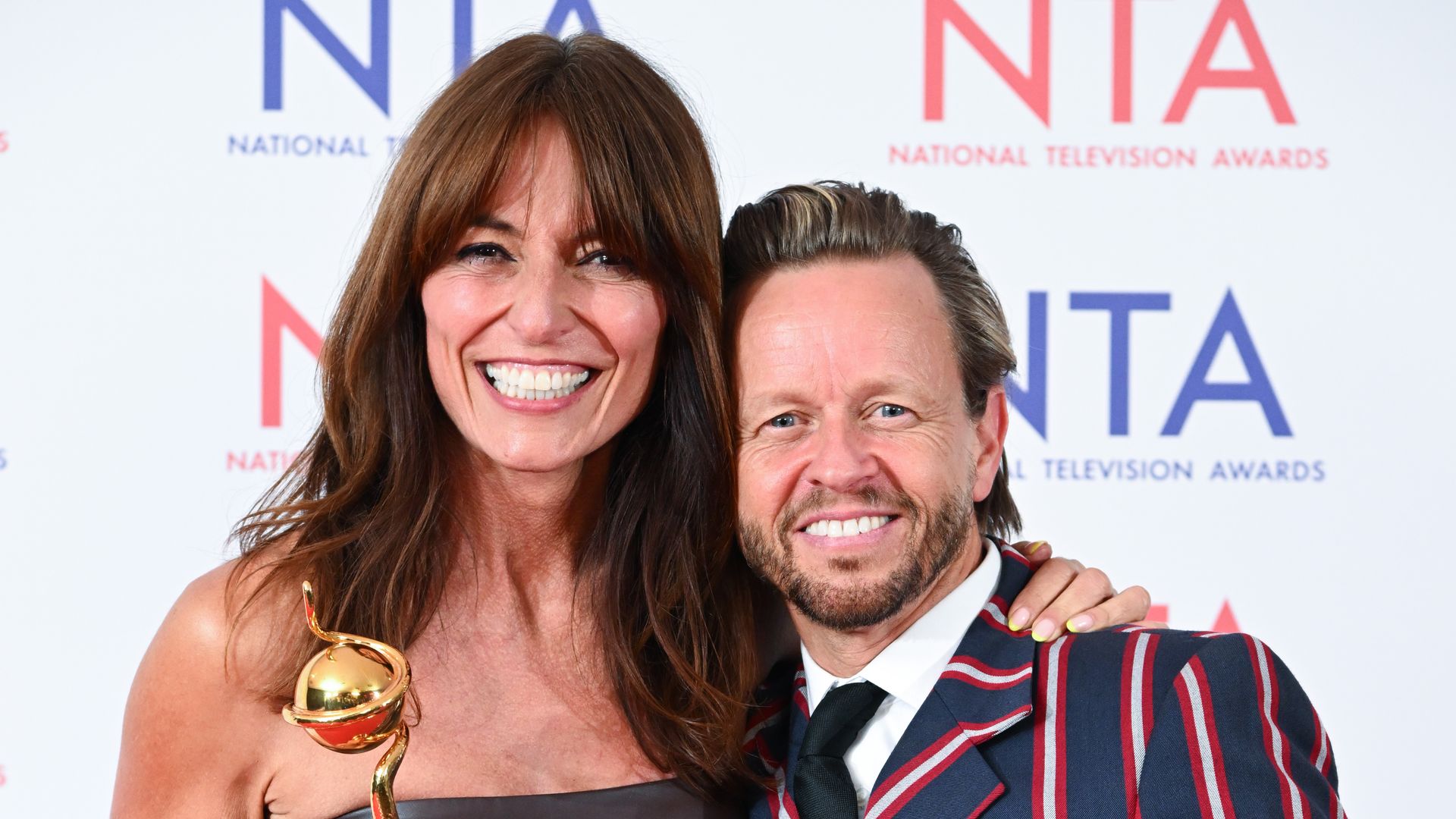 Davina McCall in a black dress holding an award with Michael Douglas in a pinstripe suit with his arm around her