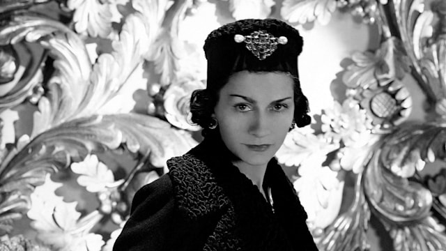 Coco Chanel wearing one of her designs: a three-quarter coat with cinched waist and astrakhan lapels and cuffs, and matching astrakhan hat with large brooch on front. 