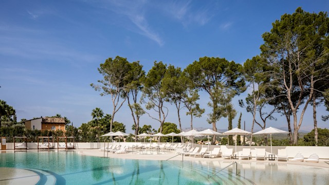 A serene poolside view at the Kimpton Aysla Mallorca in Spain, featuring crystal clear waters, white sun loungers under elegant umbrellas, and lush green trees under a bright blue sky.