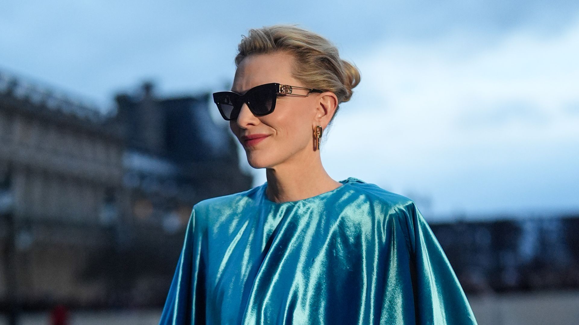 Cate Blanchett wears sunglasses, earrings, a blue turquoise gathered sleeveless lustrous shiny silk top, a bracelet, black leather flared pants, outside Louis Vuitton