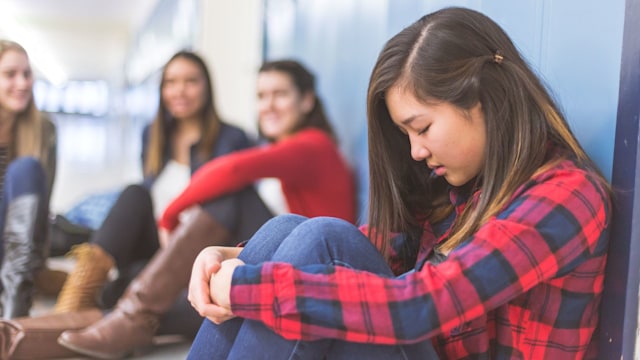 high school student leans against a panel of lockers with her eyes closed, head down, and hands clasped to her knees. Three girls are in the background talking amongst themselves and laughing at her.