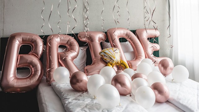 Beautiful balloons on a bed in a hotel room spelling the word bride. Beautiful decorations for a bachelorette party. The balloons are white and pink. The room is minimalistic.