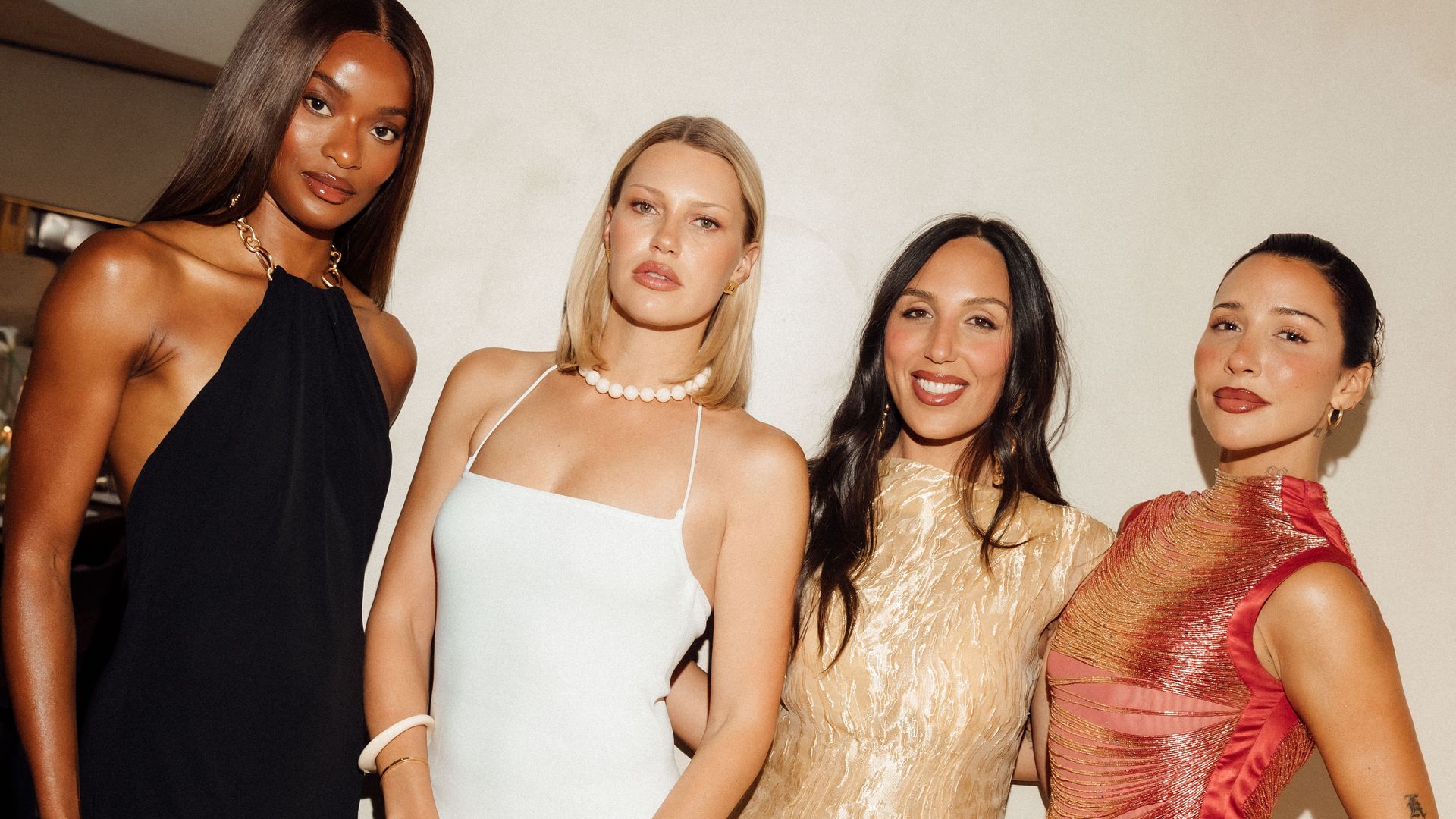 Four impeccably dressed women pose against a minimal cream-toned wall at the Cult Gaia dinner in Notting Hill, each in a distinct evening look—sleek black halter, white column dress, gold metallic gown, and a shimmering red mini.