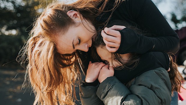 Young girl grips her mothers arm tightly and hides in the crook of her arm. Her mother gently strokes her head and talks into her ear.