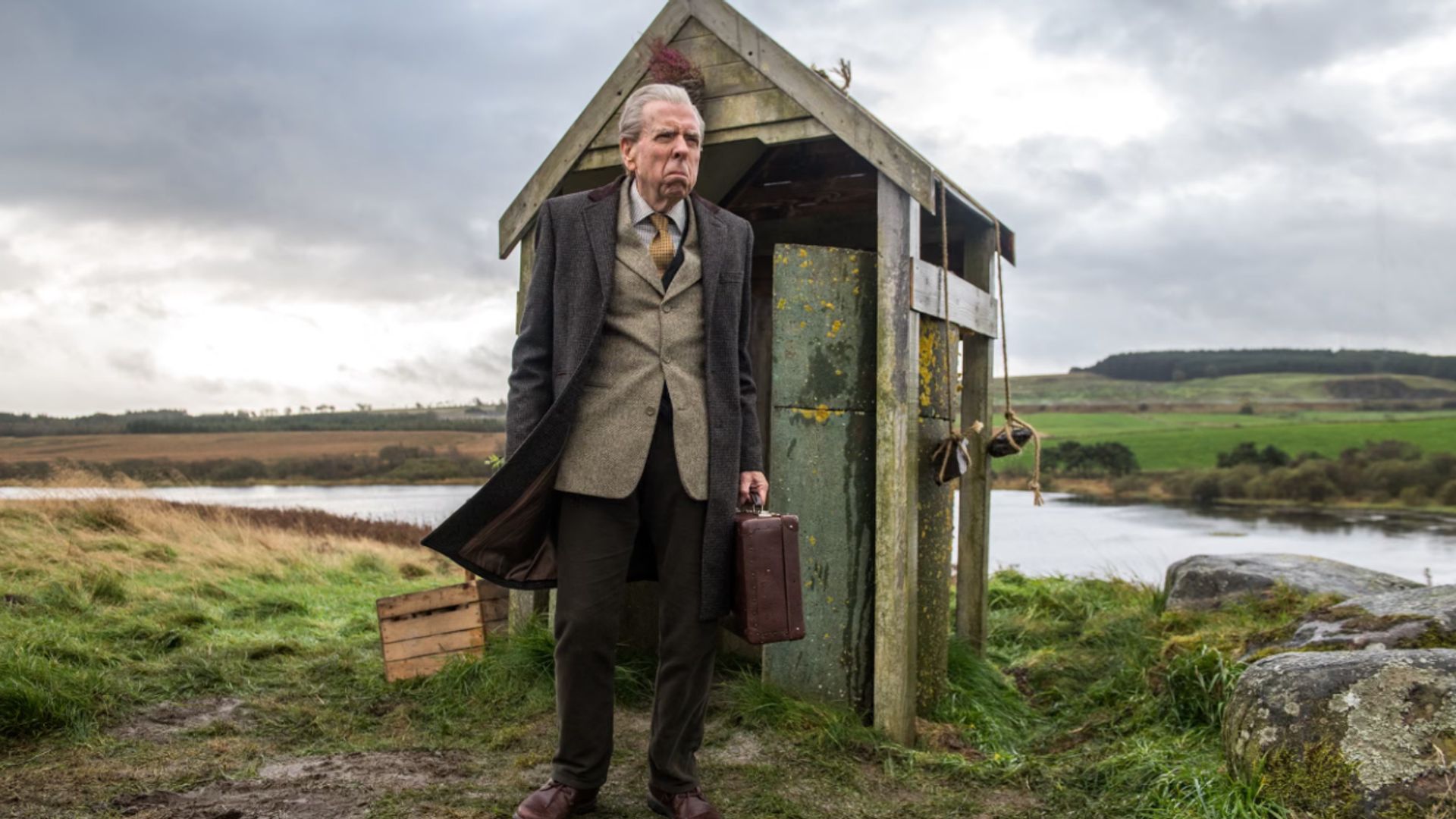 Timothy Spall standing outside a run-down bus shelter in The Last Bus