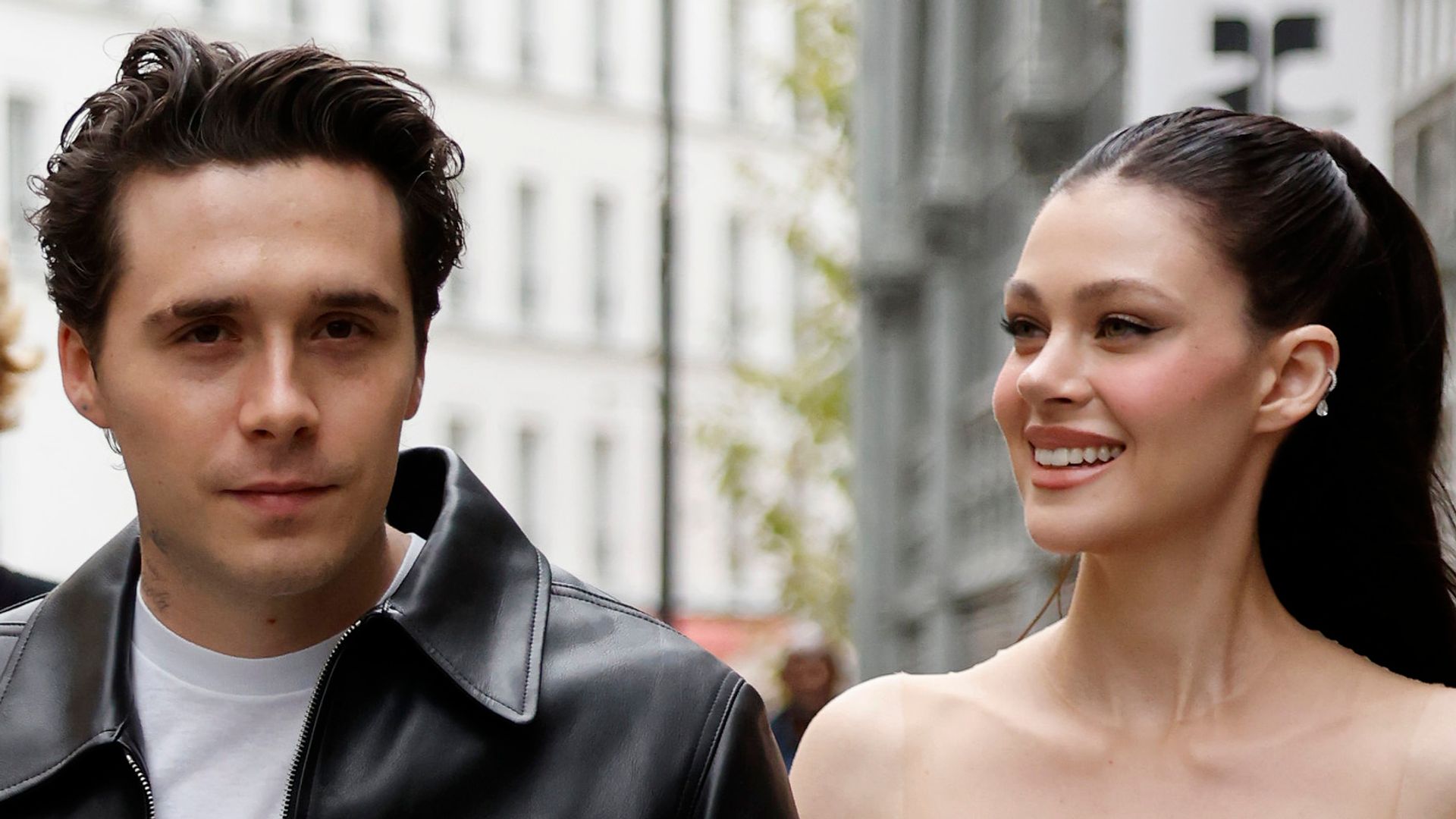 Brooklyn Beckham wears white t-shirt, black leather jacket, matching pants, black boots, and Nicola Peltz wears white jumpsuit with sheer details walking in paris street