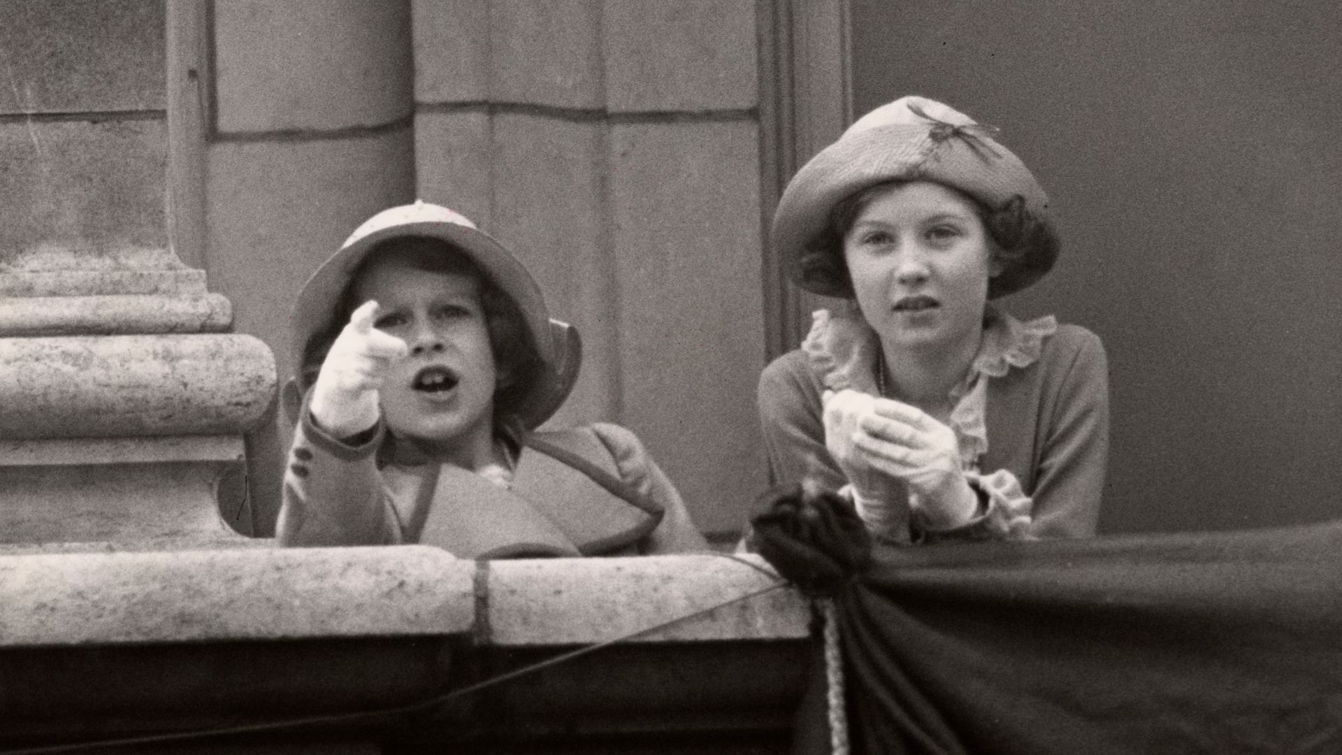Black-and-white photo of a young Queen and girl pointing out over Buckingham Palace