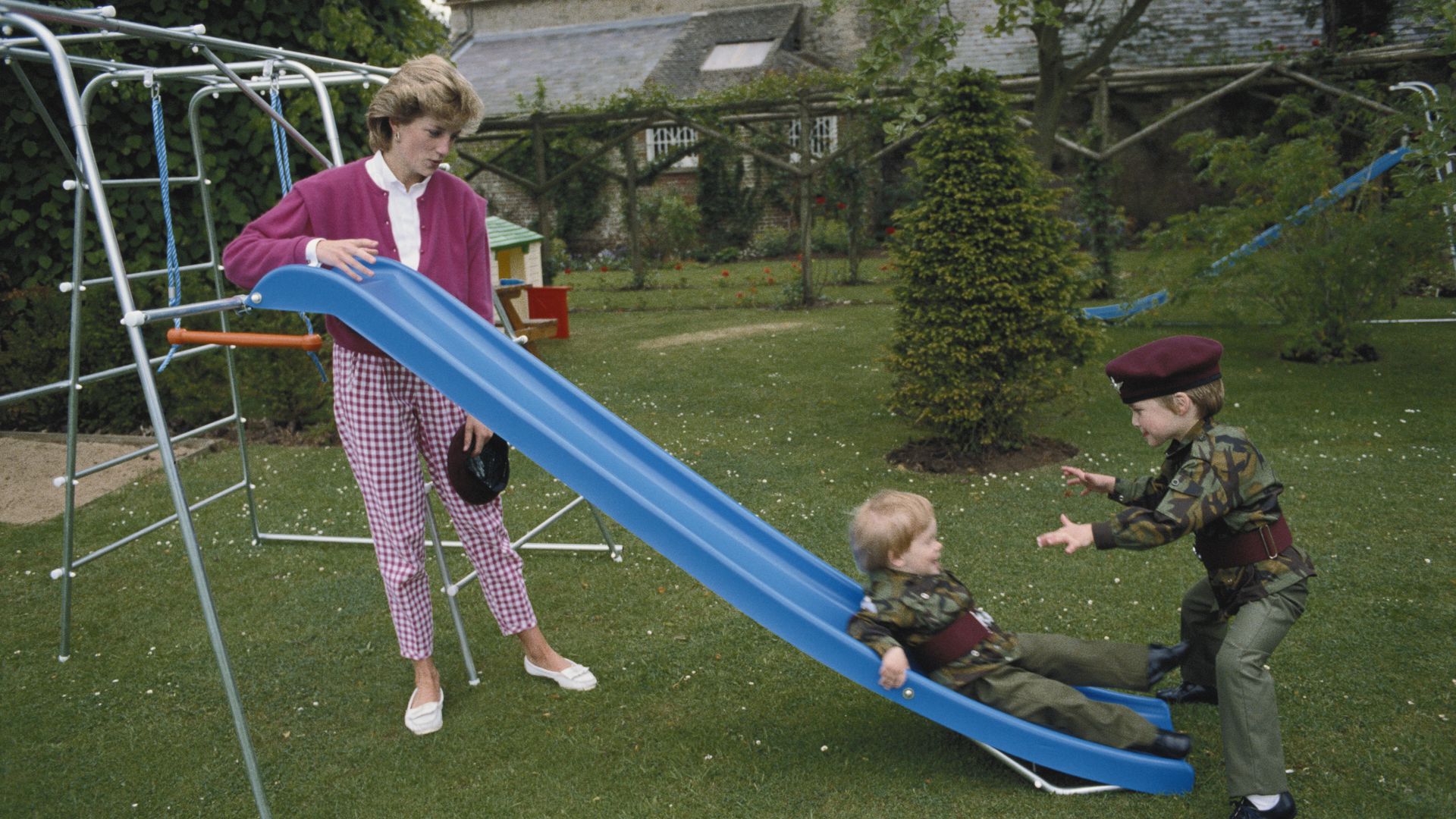 A young Prince Harry going down a slide towards Prince William; Princess Diana stands behind the slide
