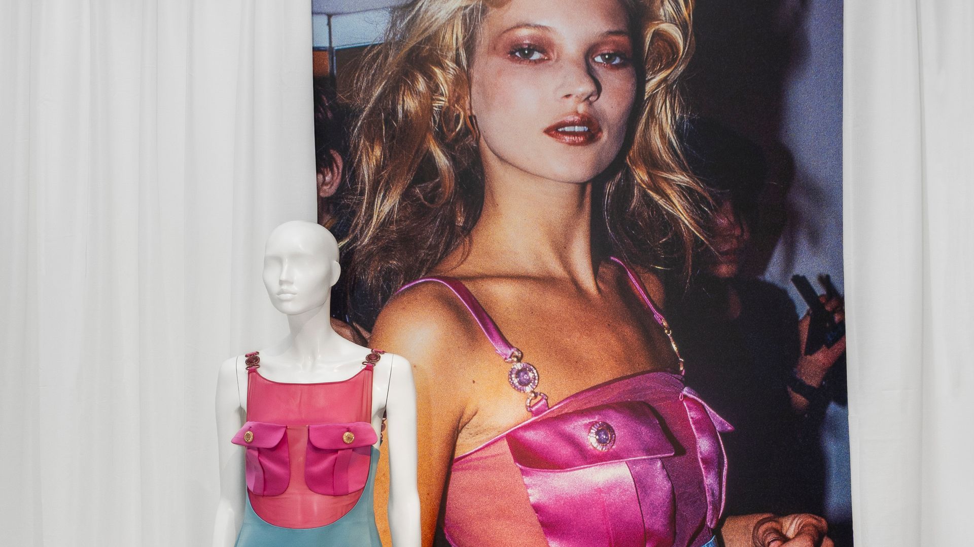A mannequin displays a colour-blocked satin mini dress from Gianni Versace, worn by Kate Moss, featuring a hot pink bustier top and aqua blue skirt, set against a large photograph of Moss wearing the original look backstage in the '90s.