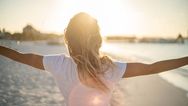 Cheerful young woman embracing nature at sunset; female standing on beach arms outstretched, Cancun, Mexico