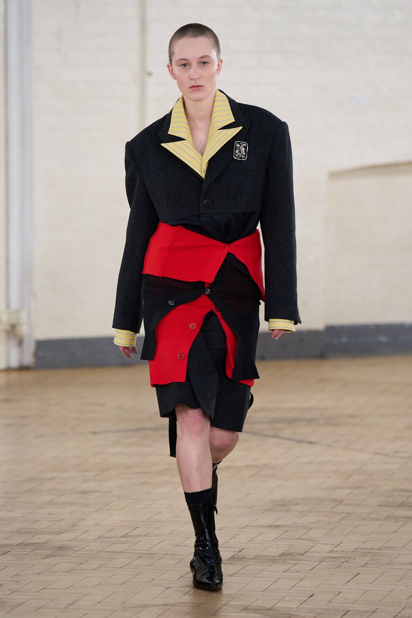 Du drame vintage au nouveau minimalisme, les plus grandes tendances de LFW AW26 15 Un mannequin porte un blazer noir avec un revers jaune et un tricot rouge enroulé autour de sa taille, une jupe noire, des chaussettes noires et des chaussures noires.