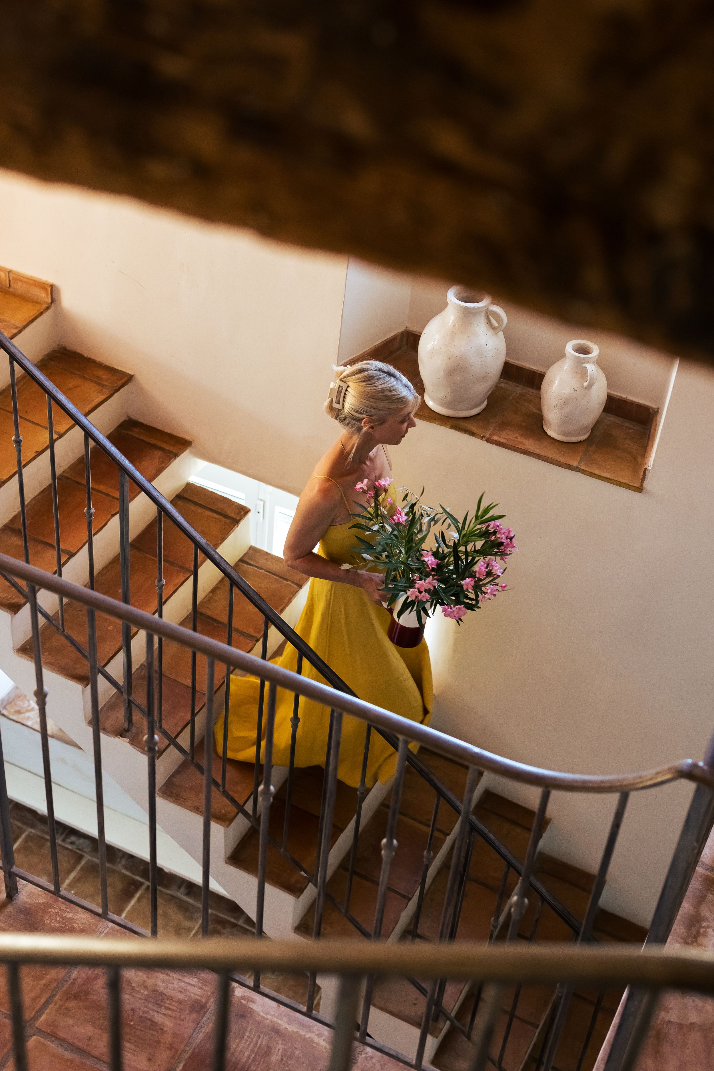 Jeany walking down the stairs in a yellow dress holding pink flowers in Domaine Mirabeau Provençal manor house