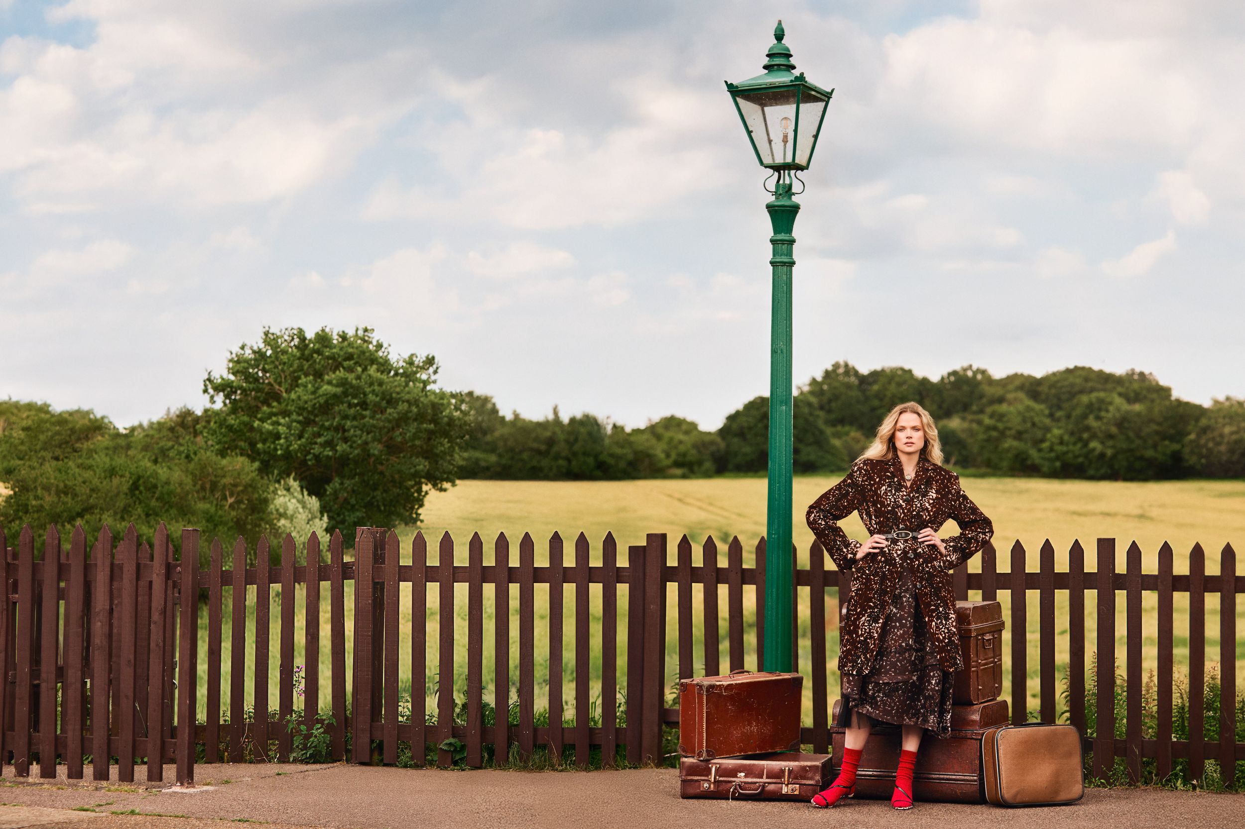 Gabriella Wilde stands at a rural train platform in a belted leopard-print coat, surrounded by vintage luggage, with a wide stance and vibrant red socks stealing the show.