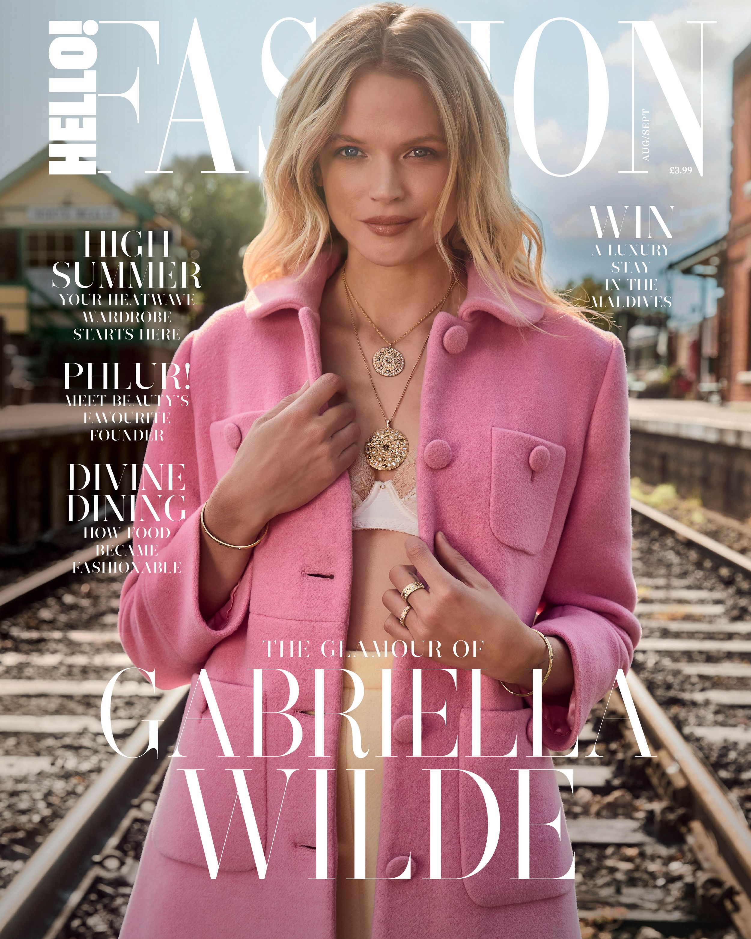 Gabriella Wilde stars on the August–September 2025 cover of Hello! Fashion, wearing a bubblegum pink coat over a bralette and skirt, layered with gold medallion necklaces from De Beers and styled against sunlit railway tracks.