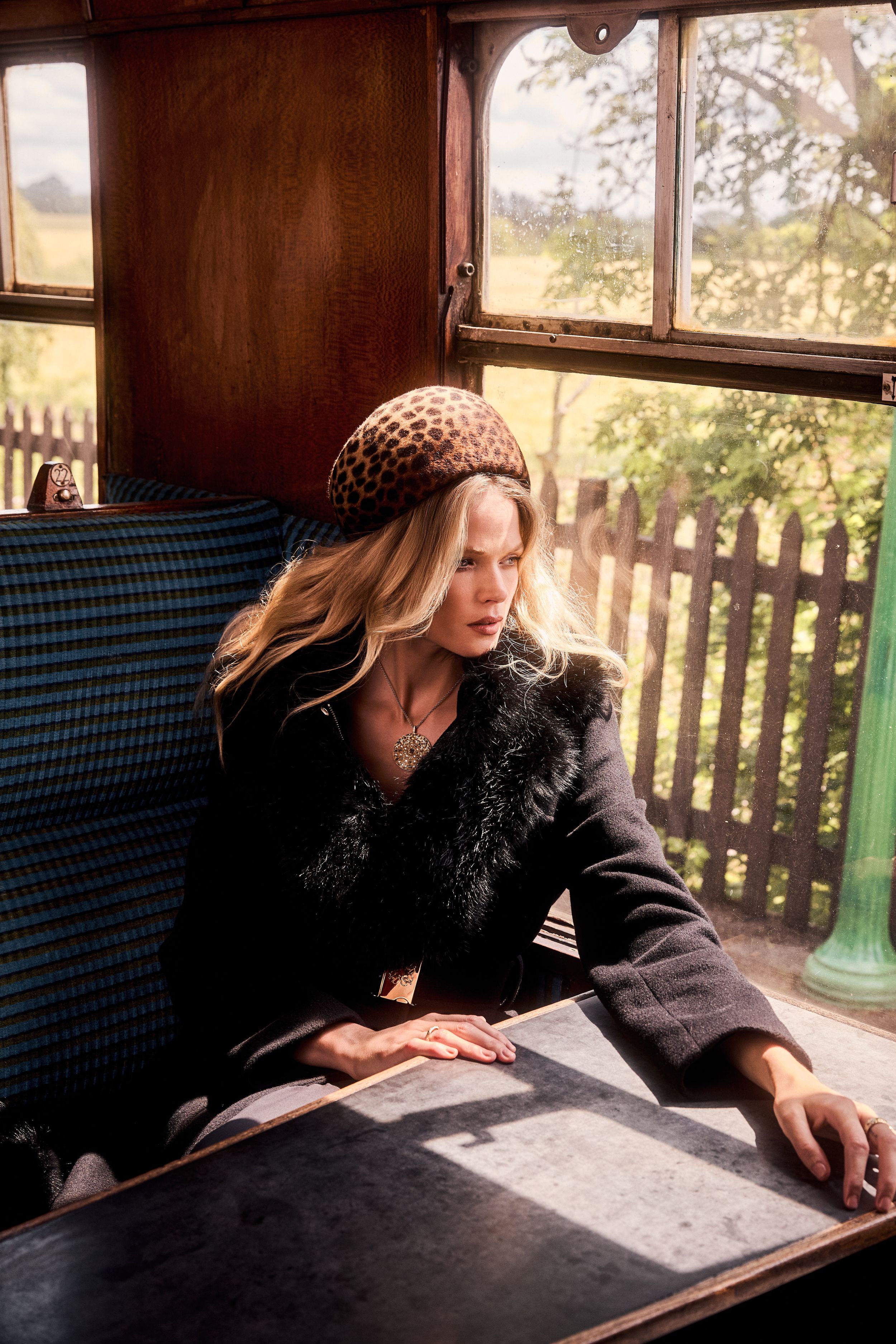 Gabriella Wilde gazes out of a train window in a leopard beret and black coat with a dramatic faux fur collar, paired with a statement pendant necklace and windswept curls.