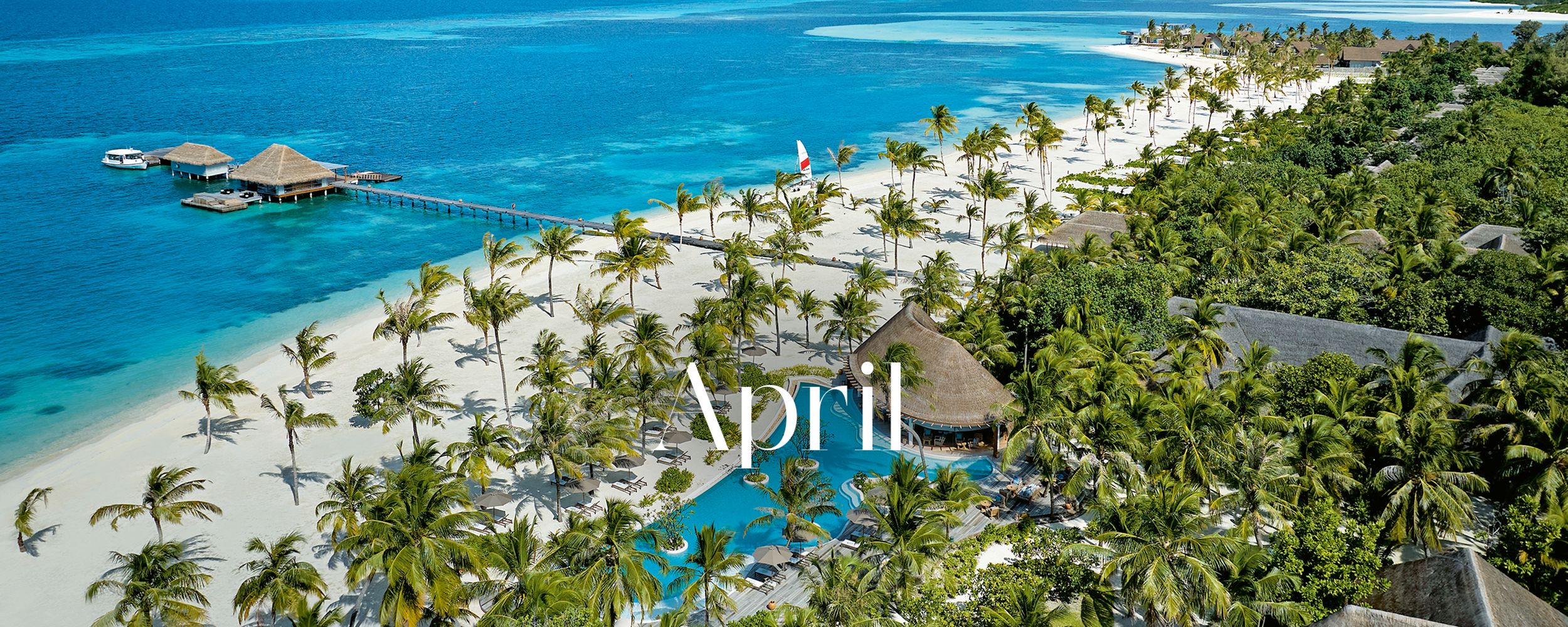 Aerial view of a tropical beach resort with turquoise waters, a wooden pier, palm trees, and a pool